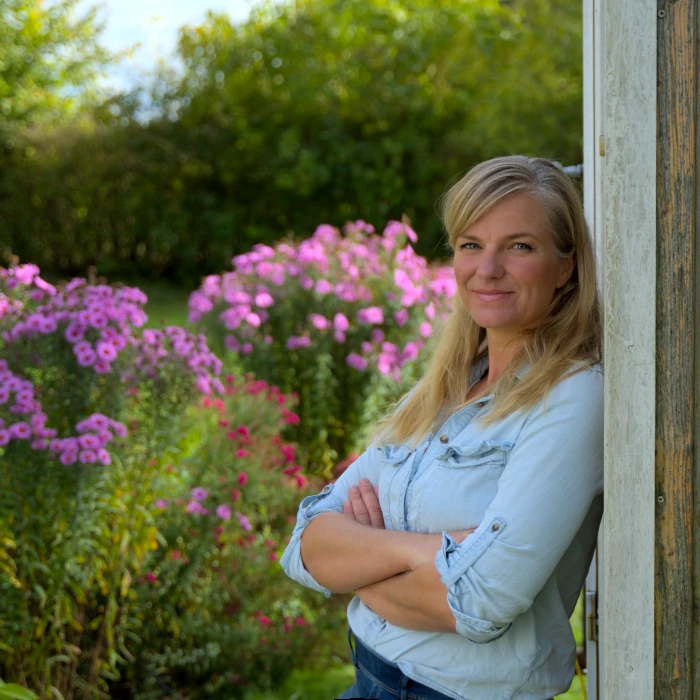 Verena mit hellblauer Bluse in ihrem Garten, im Hintergrund lila blühende Astern.